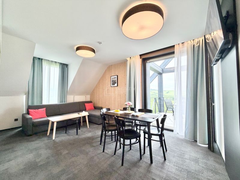 Svetlá obývacia izba v rekreačnom apartmáne s balkónom, sivou sedačkou a jedálenským stolom.
