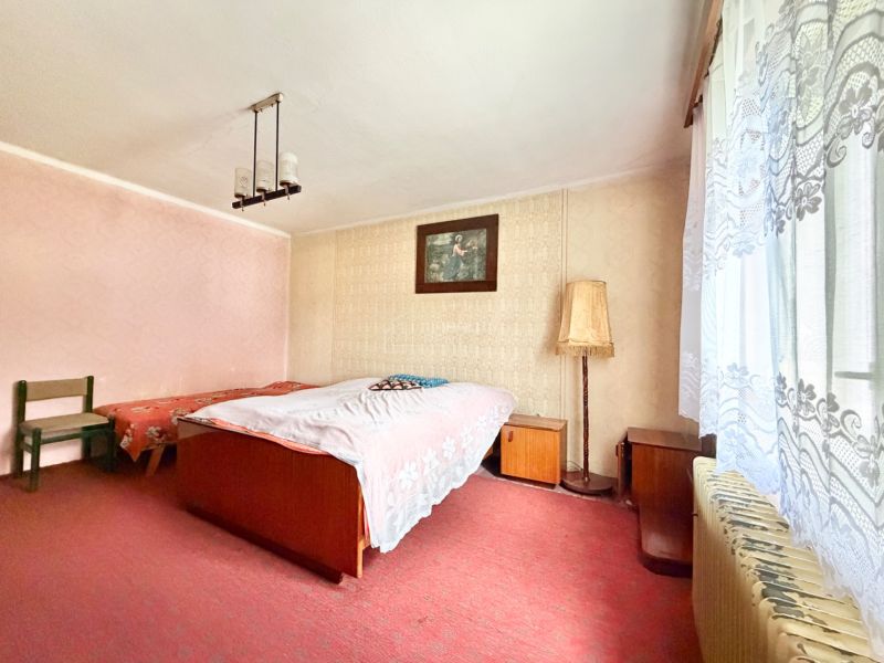 Spálňa v rodinnom dome, svetlé tapety, koberec, vintage nábytok a veľké okno s čipkovanými záclonami.